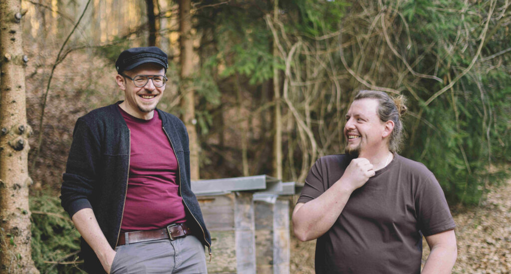 Es sind Richard und Michael mit zwei Bienenstöcken im Hintergrund zu sehen. 